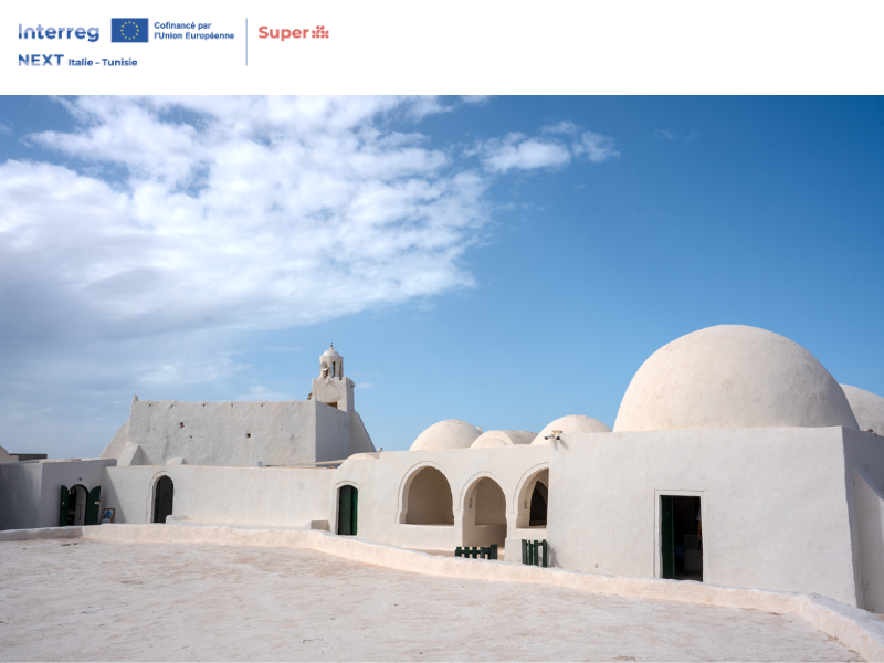 Veduta dell’architettura tradizionale di Djerba, in Tunisia, con edifici bianchi dalle cupole arrotondate e archi, sotto un cielo azzurro. L’immagine riporta i loghi del progetto SUPER – Interreg NEXT Italie-Tunisie.